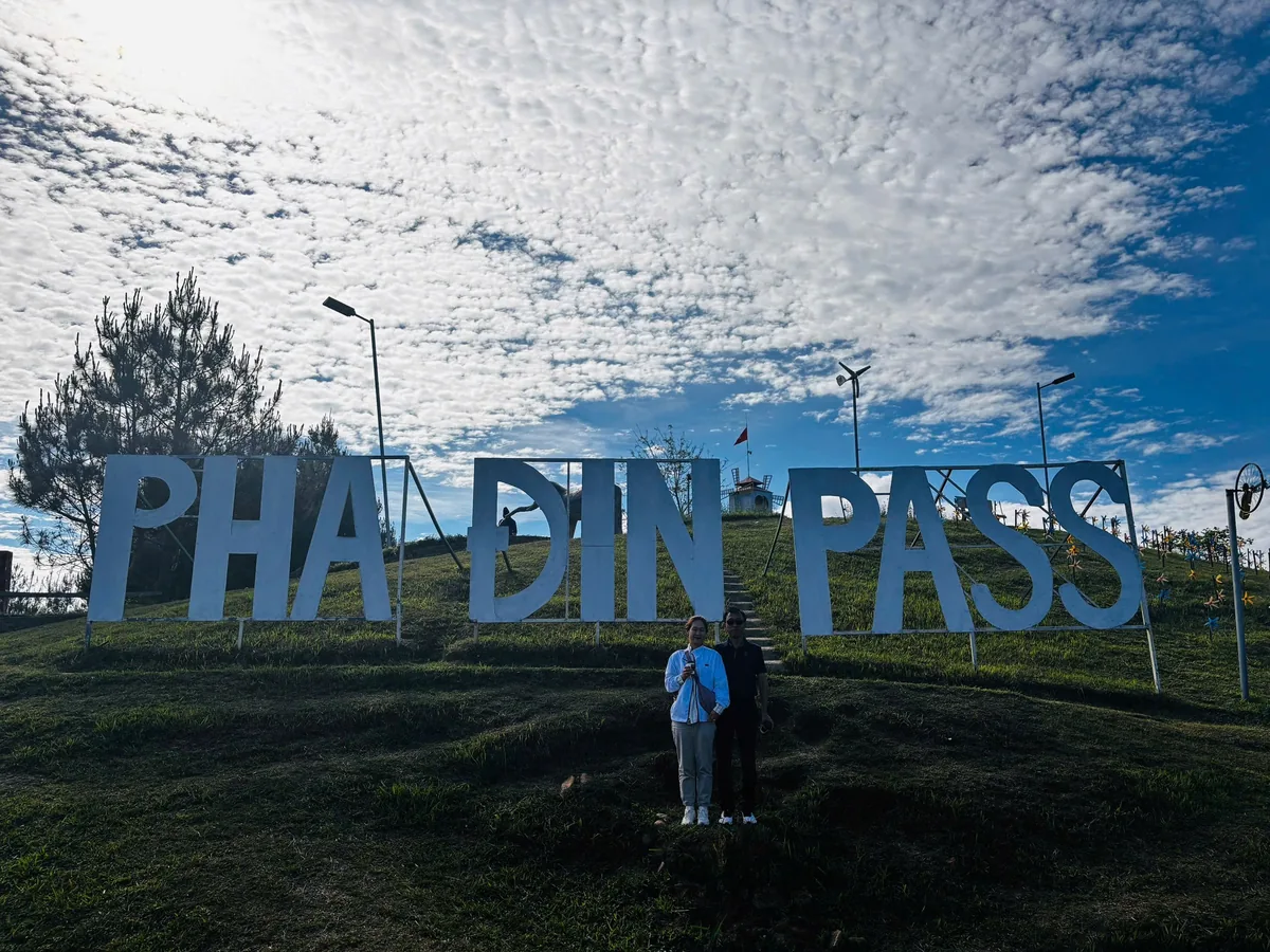 Hai du khách chụp ảnh dưới biển chữ trắng lớn Pha Din Pass trên đồi cỏ dưới bầu trời nhiều mây xanh.